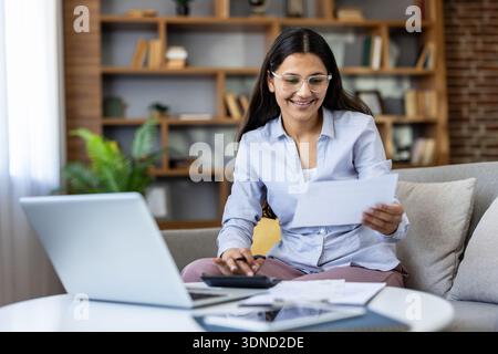 Junge Frau lächelt, während sie zu Hause sitzt, Dokumente sorgfältig überprüft, Taschenrechner und Laptop benutzt, persönliche Finanzen verwaltet und Steuern vorbereitet Stockfoto