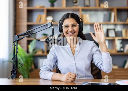 Junge lächelnde Frau mit Kopfhörern und Brillen, die Podcasts oder Livestreams aufnimmt, mit einem professionellen Mikrofon an einem Schreibtisch sitzt und dem Publikum eine Hand zuwinkt Stockfoto