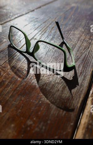Ein gebrochenes Paar grüner Gläser mit fehlendem Arm liegt auf einem sonnendurchfluteten Holztisch, was Verlust und Unvollkommenheit hervorhebt. Stockfoto