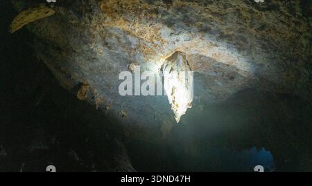 Dramatischer Blick in eine Höhle mit einer hellen Taschenlampe, die einen großen Stalaktiten beleuchtet, der von der felsigen Decke hängt, mit dunklen Schatten und rauem Text Stockfoto