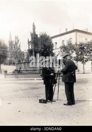 Zwei Männer stehen neben einer auf einem Stativ montierten Kamera in einem offenen Quadrat, einer hält Papiere, der andere passt die Ausrüstung an. Hinter ihnen erhebt sich ein kunstvoller Brunnen mit Skulpturen, eingerahmt von Bäumen und fernen Gebäuden. Die Szene fängt einen Moment der Vorbereitung in dieser Ansicht von Clermont-Ferrand aus dem Jahr 1927 ein. Aus einer Sammlung einzigartiger Fotografien des 20. Jahrhunderts. Amateurfotograf, Schnappschüsse und einige größere in einem einzigen Album, das die Reisen einer Familie in Europa dokumentiert. Stockfoto