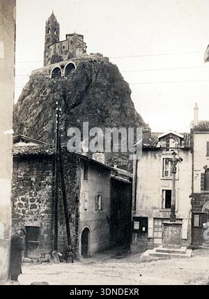 Ein steiler vulkanischer Gipfel in Le Puy‑en‑Velay erhebt sich über Häusern, die von der Kapelle Saint‑Michel gekrönt werden. Der hohe romanische Turm und das kompakte Heiligtum stehen dramatisch auf dem Felsen. Enge Gassen, gekachelte Dächer und Versorgungsmasten füllen den Vordergrund und setzen den Alltag des Dorfes mit dem markanten Hochdenkmal in Kontrast. Aus einer Sammlung einzigartiger Fotografien des 20. Jahrhunderts. Amateurfotograf, Schnappschüsse und einige größere in einem einzigen Album, das die Reisen einer Familie in Europa dokumentiert. Stockfoto