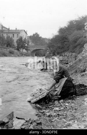 Eine Frau kniet auf dem felsigen Uferufer und arbeitet über eine flache Oberfläche mit einem Tuch, das neben ihr verteilt ist. Eine andere Frau steht weiter flussaufwärts, die ebenfalls mit dem Waschen beschäftigt ist. Gebäude, Bäume und eine Steinbrücke säumen das gegenüberliegende Ufer und verankern die Szene in einer ruhigen Ecke am Fluss der Altstadt. Aus einer Sammlung einzigartiger Fotografien des 20. Jahrhunderts. Amateurfotograf, Schnappschüsse und einige größere in einem einzigen Album, das die Reisen einer Familie in Europa dokumentiert. Stockfoto