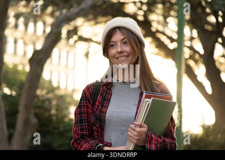 Eine Schülerin lächelt, hält einen grünen Notizblock, trägt Mütze und Rucksack auf dem Campus Stockfoto