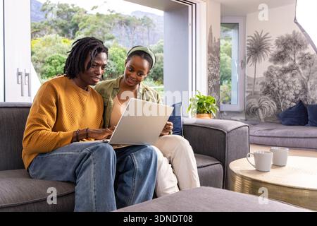 Afroamerikanisches Paar, das auf einem grauen Sofa in einem hellen Wohnzimmer sitzt und sich einen silbernen Laptop teilt Stockfoto