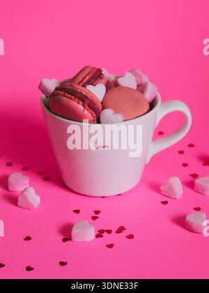 Liebe in einer Tasse - Valentinstag-Karte Idee. Rosafarbene Makronen und Zuckerherzen in einer Tasse mit geschriebenem Wort LIEBE auf rosa Hintergrund Stockfoto