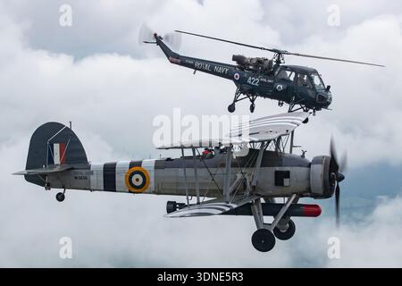 Nahaufnahme eines Fairey-Schwertfisches Mk I, der 2024 mit einem Westland Wasp HAS.1 über Sleapkosh, Shropshire, in Formation fliegt. Beide Flugzeuge tragen die Royal Navy-Ausstellungspläne. Der Swordfish Mk I ist ein dreisitziger, einmotoriger zweiflügeliger Torpedobomber, der von einem luftgekühlten Bristol Pegasus IIIM-9-Zylinder-Radialkolbenmotor angetrieben wird. Der Westland Wasp HAS.1 ist ein kompakter Marinehubschrauber, der von einem Bristol Siddeley Nimbus Turbowellenmotor angetrieben wird und einen vierblättrigen Hauptrotor antreibt. Das Foto betont Cockpitbereiche, Rumpfdetails und koordinierten Formationsflug. Stockfoto