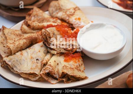 Dünne Crepes, die auf einem Teller mit rotem Kaviar und Sauerrahm gefaltet sind, werden als traditionelles festliches Frühstück oder Vorspeise serviert. Stockfoto