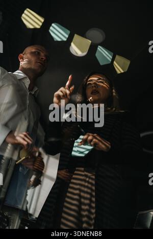 Zwei Kollegen ziehen ein Brainstorming unter dekorativen Bannern durch und platzieren während einer gemeinsamen Bürositzung Haftnotizen Stockfoto