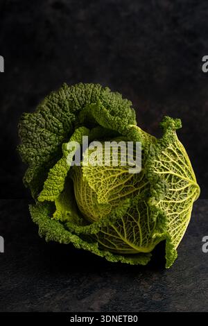 Ganzer wirsingkohl auf dunklem, strukturiertem Hintergrund, dramatisches natürliches Licht, das seine knittergrünen Blätter hervorhebt. Stockfoto