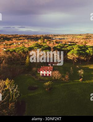 Aus der Vogelperspektive auf ein einsames rot-weißes Haus eingebettet zwischen den üppigen grünen Feldern und den dichten Wäldern unter einem Himmel, der mit Dämmertönen bemalt ist Stockfoto