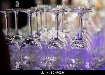 Auf dem Kopf stehende Weingläser in ordentlichen Reihen mit violetter und blauer Veranstaltungsbeleuchtung, glitzernden Reflexen und weichem Bokeh-Hintergrund im Barservice-Bereich Stockfoto