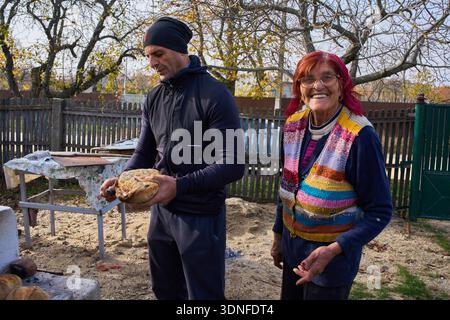 Mann mittleren Alters, der frisch gebackenes Brotbrot neben lächelnder älterer Mutter im ländlichen Hinterhof hält Stockfoto