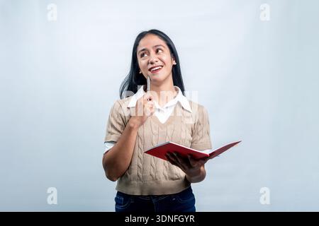 Eine glückliche junge indonesische Frau, die ein offenes rotes Notizbuch und einen Stift hält und nachdenklich aufblickt, während sie träumt oder ein kreatives Projekt plant. Stockfoto