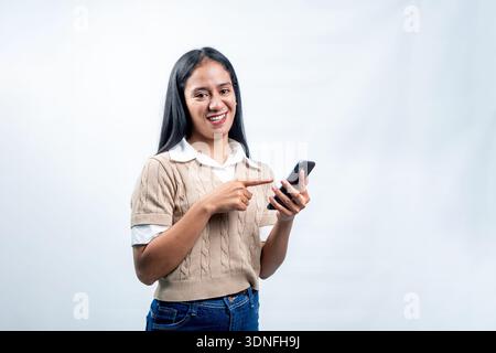 Eine glückliche junge indonesische Frau, die ein Mobiltelefon hält und auf den Bildschirm zeigt, eine App, eine Website oder digitale Inhalte mit einem freundlichen Lächeln präsentiert Stockfoto