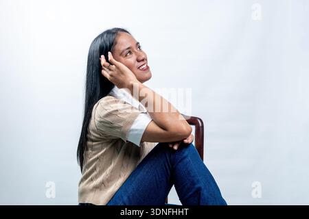 Fröhliche junge indonesische Frau in lässigem, elegantem Outfit, die auf einem Stuhl sitzt. Seitliches Porträt mit einem freundlichen Lächeln auf einem sauberen weißen Hintergrund. Stockfoto