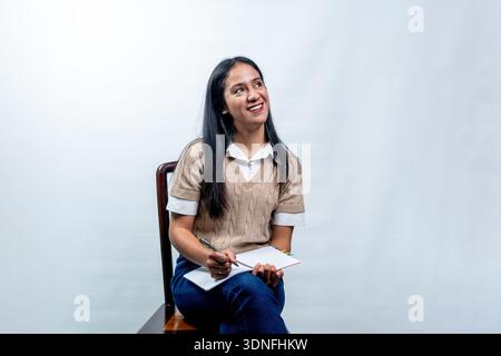 Eine glückliche junge asiatische Frau mit langen schwarzen Haaren, die auf einem Stuhl sitzt und mit einem hellen Lächeln über die Schulter blickt. Stockfoto