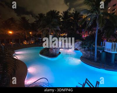 NUR REDAKTIONELLE NUTZUNG - MARGARITAVILLE RESORT HOLLYWOOD BEACH FLORIDA: Ein Nachtblick auf den Lagunenstil Swimmingpool im Margaritaville Hollywood Beach Resort, mit einem prominenten künstlichen Felsen Wasserfall und tropischer Landschaft. Der Pool ist mit leuchtend blauer und rosafarbener Unterwasser-LED-Beleuchtung beleuchtet, mit den mehrstöckigen Flügeln des Resorts und dunklen Palmensilhouetten im Hintergrund. Dieses Erholungsgebiet ist eine der wichtigsten Annehmlichkeiten für das Resort am Hollywood Beach Broadwalk. Stockfoto