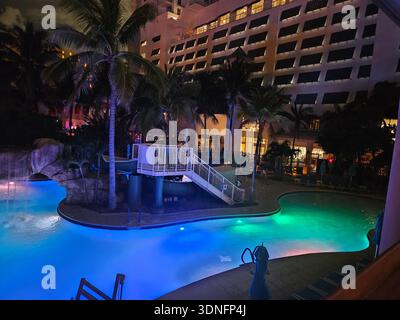 NUR REDAKTIONELLE NUTZUNG - MARGARITAVILLE RESORT HOLLYWOOD BEACH FLORIDA: Ein abendlicher Blick auf den beleuchteten Pool im Lagunenstil im Margaritaville Hollywood Beach Resort. Die Szene bietet eine leuchtend blaue und grüne Unterwasserbeleuchtung, einen Felswasserfall und eine Wasserrutsche vor dem Hintergrund der gestuften architektonischen Balkone und tropischen Palmen des Resorts. Diese reizvolle Gegend ist ein Herzstück des Reiseziels mit 349 Zimmern am Hollywood Beach Broadwalk. Stockfoto