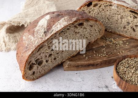 Frisch gebackenes und in Scheiben geschnittenes Brot aus Dinkelsauerteig mit zerstoßenen Hanfsamen ist mit Cannabisblättern aus Mehl auf Holzbrett mit Leinen verziert Stockfoto