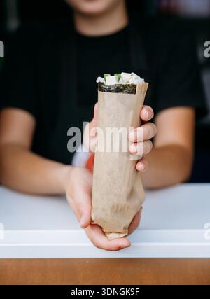 Hand hält Sushi-Burrito in Papierfolie, Straßenessen zum Mitnehmen aus der Nähe Stockfoto