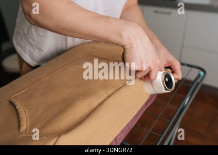 Männliche Hände verwenden Akkufusselentferner auf braunem Pullover, entfernen Fuzz und Pilling, um die glatte Textur wiederherzustellen und getragene Strickwaren für den Alltag aufzufrischen Stockfoto
