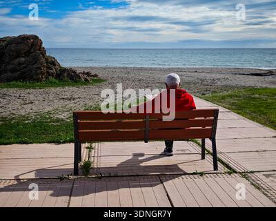 Benalmádena, Málaga, Spanien. 8. Februar 2026. Ein älterer Mann mit rotem Pullover sitzt auf einer Bank vor dem Strand in Benalmádena, Spanien. Stockfoto