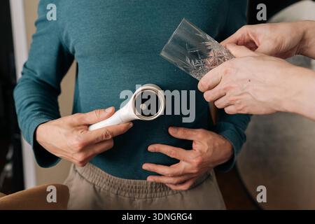 Person, die elektrischen Fusselentferner hält, das Bekleidungspflegegerät auf einem blauen Pullover bedient, um die Textiloberfläche zu glätten und Pilling zu entfernen, wodurch das Textil verbessert wird Stockfoto