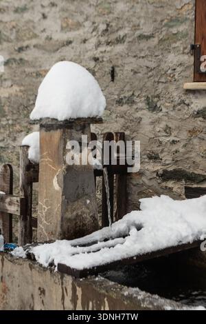 Ein rustikaler Steinbrunnen liegt unter frischem Schnee, wo klares Wasser immer wieder fließt und die Winterstille, den ländlichen Charme und die ruhige Beharrlichkeit einfängt Stockfoto