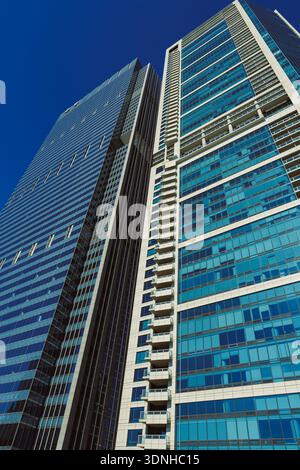 Zwei hohe Gebäude sind aus einem niedrigen Winkel dargestellt, die Sonnenlicht auf ihren Glasflächen reflektieren. Der Himmel ist klar und blau, was die Struktur hervorhebt Stockfoto
