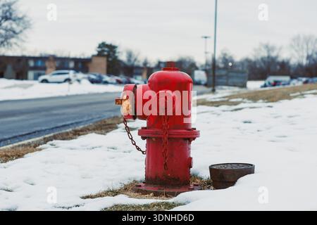 Ein roter Hydrant befindet sich am Rand eines verschneiten Bereichs neben einer Straße. Autos parken im Hintergrund unter bewölktem Himmel. Der Boden ist bedeckt mit Stockfoto