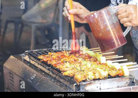 Nahaufnahme einer Handputzsoße auf Hühnerspießen auf einem Nachtmarkt. Authentische Street Food-Zubereitung und Outdoor-Cooking-Action-Szene. Stockfoto