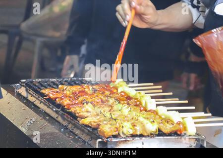 Nachtmarktverkäufer bürsten Grillsoße auf Hähnchenspieße über offenem Grill. Authentische Street-Cooking-Atmosphäre. Stockfoto