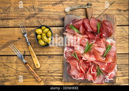 Erstklassiges Trockenfleisch auf rustikalem Holz: Schinken, coppa und Bresaola garniert mit Rosmarin und grünen Oliven, Überkopf. Stockfoto