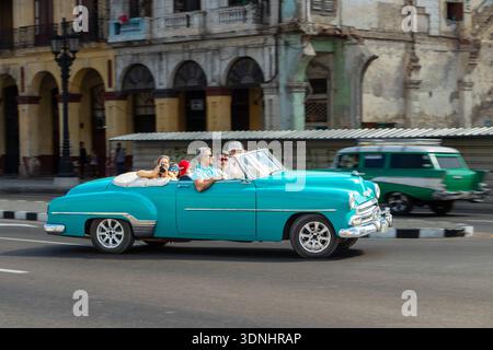 Touristen genießen eine Fahrt in einem klassischen, türkisfarbenen amerikanischen Cabrio, das in einer historischen Stadt eine Straße hinunter fährt Stockfoto