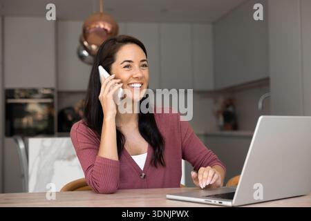 Lächelnde Frau, die am Telefon spricht, am Tisch mit Laptop sitzt Stockfoto