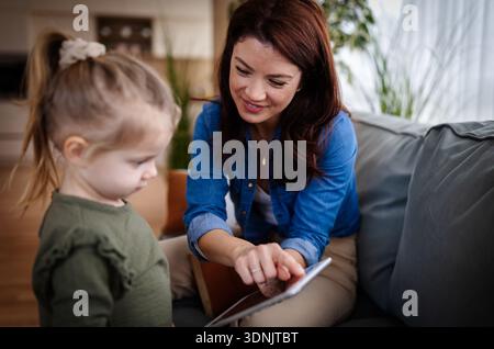 Eine Mutter zeigt ihrer Tochter eine Tafel, während sie auf einer Couch sitzt. Sie befinden sich in einem hellen Wohnzimmer mit Pflanzen und einer warmen Atmosphäre. Stockfoto