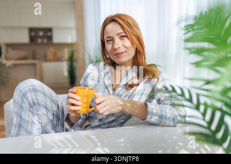 Eine Frau entspannt sich auf einer Couch, die einen gelben Becher hält, während sie einen Pyjama trägt. Sonnenlicht wird durch das Fenster gefiltert und schafft eine warme Atmosphäre. Stockfoto