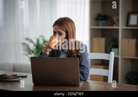 Eine Person sitzt an einem Tisch mit offenem Laptop vor sich. Sie trinkt aus einer Tasse und konzentriert sich auf ihre Arbeit. Cookies sind in der Nähe zu finden. Stockfoto