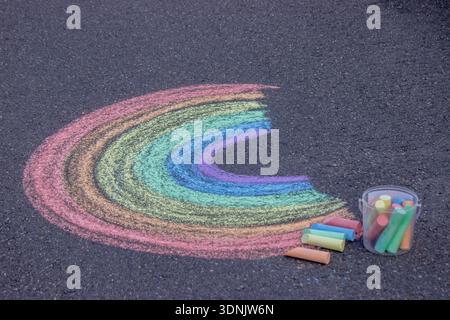 Farbenfroher Regenbogen, gezeichnet mit Kreide auf Asphaltpflaster mit einem Eimer Buntstifte. Stockfoto
