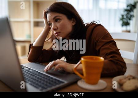 Eine Frau sitzt an einem Tisch im Wohnzimmer. Sie konzentriert sich auf ihren Laptop, während sie Tee schlürft. Sie fühlt sich müde und gelangweilt Stockfoto