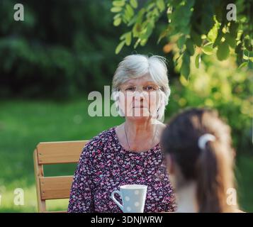 Ältere Frau, die sich mit ihrer Enkelin draußen an einem schönen sonnigen Tag hinsetzt, Tee genießt und ein Gespräch führt Stockfoto
