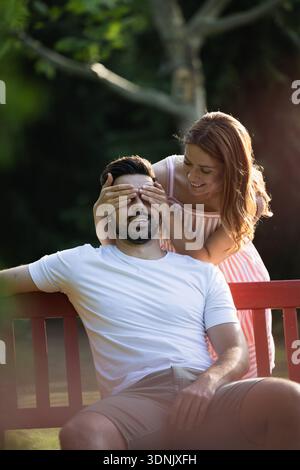 Frau überrascht und deckt die Augen des Freundes an einem sonnigen Tag im Park. Der glückliche Mann, der auf einer Bank sitzt Stockfoto
