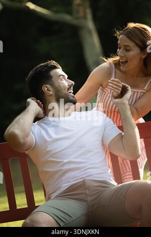 Frau überrascht Freund von hinten, während Mann an einem sonnigen Tag auf einer Bank im Park sitzt Stockfoto