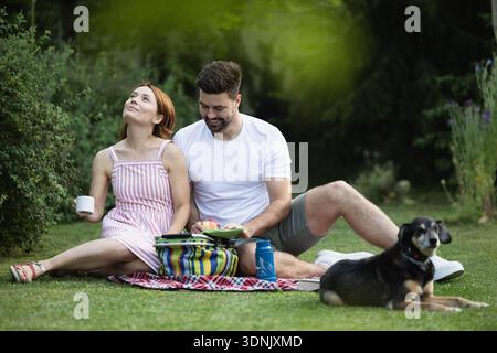 Junge Paare genießen ihr Picknick, essen und trinken, während sie zusammen mit ihrem Hund in der Natur sitzen Stockfoto