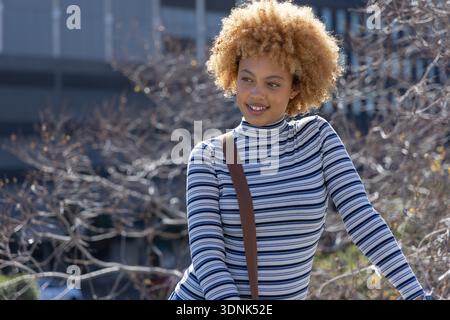 Afroamerikanerin steht und lächelt, trägt gestreiftes Top und braunes Crossbody am Urban plaza Stockfoto