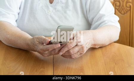 Handy in den Händen einer älteren Frau auf Holztischhintergrund. Ältere Frau, die Handy in der Hand hält und kippt. Kommunikation alter Menschen Stockfoto