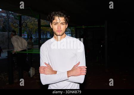 Porträt eines jungen Mannes mit lockigen Haaren in Sportkleidung, der in der Abenddämmerung in einem urbanen Outdoor-Fitnessstudio steht Stockfoto