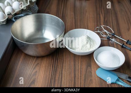 Backzutaten, die vor dem Mischen angeordnet sind, einschließlich Mehl, Zucker, Eier und Schüsseln auf einer hölzernen Küchenarbeitsfläche. Stockfoto