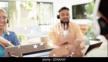 Führender bärtiger Mann in orangefarbenem Hemd, der im Büro zuhört, mit Laptops, Kopfhörern, Sterngrafiken Stockfoto
